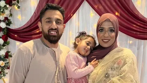 Akeel Nanabawa and Hannaa Vorajee hold their daughter Sara at a celebration event. Akeel wears a grey top while Hanaa wears a terracota head scarf and gold top. Sara is dressed in pink and they stand in front of a floral display