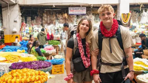BBC / Studio Lambert Fin and Sioned pictured at a market in India. They are wearing a traditional Indian flower necklace, known as mala or haar. They also both have floral bracelets on which match the necklace. They are both smiling at the camera and wearing backpacks.
