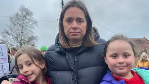 A woman in a black puffer coat stands with her arms round two girls. Other protesters can be seen behind them.