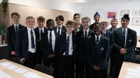 BBC A photo of a group of teenagers standing and smiling into the camera they are all wearing a school uniform. 