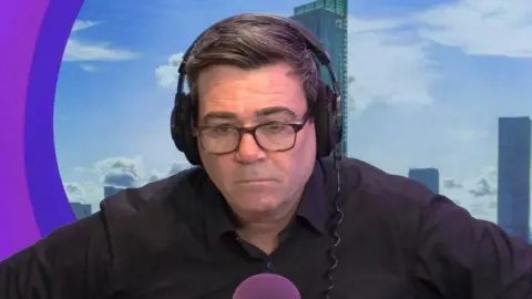 Andy Burnham wearing a black shirt and black headphones sits above a BBC Radio Manchester purple microphone in a radio studio. 