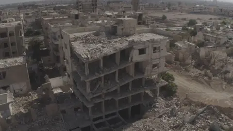A high vantage camera looks down on a large area of partially demolished and destroyed buildings. In the centre of the image is a building which is collapsing and in ruin.