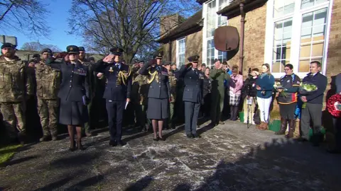 A large group of servicemen and women at the memorial in their uniforms are saluting. It is a sunny day.