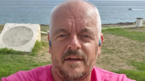 Brain Tumour Research A man with very short white hair and brown and grey facial hair is taking a selfie. He is wearing grey in-ear headphones and a pink t-shirt. There is grass in the back