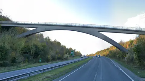 Google A footpath bridge going across the A41 near Tring. Two carriageways with a few cars in the distance. The road has trees either side. 