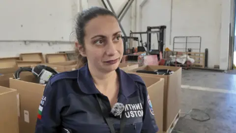 Sonia Daneva, a customs official, is standing in a customs shed at the Kapitan Andreevo border between Bulgaria and Turkey. She's standing in front of a number of boxes and a fork lift truck.
