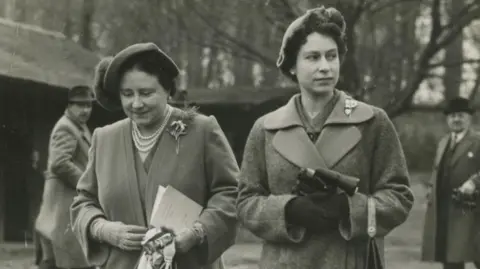 A black and white photograph of Queen Elizabeth, The Queen Mother, and Queen Elizabeth II. Both are wearing long coats and hats. 