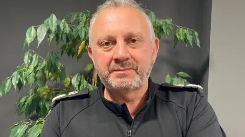 BBC A mid shot of Damian Kitchen. He has short grey hair and a grey beard. He is wearing a black shirt with police shoulder pads.