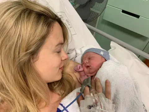 Kimberley Nixon A blonde woman lying in a hospital bed with tubes coming out of her hand. She is looking down at a tiny baby with a blue and white striped hat on in her arms.