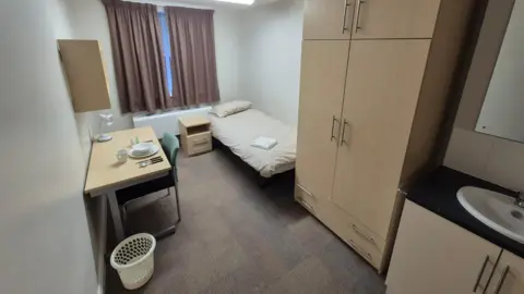 West Northamptonshire Council A room with a bed in it with cream covers. There is a desk to the left with crockery and a lamp on it. There is a wardrobe by the bed, and a sink with cupboards below it and a mirror above. There is a cream bin on the left and a green chair at the desk. There is also a bedside table.