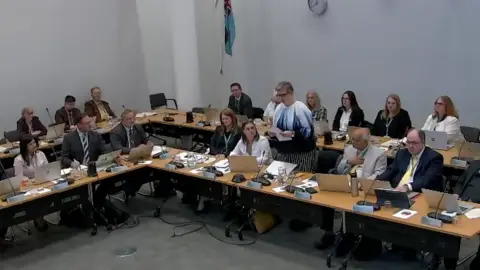 A still of Dacorum Borough Council's full meeting where many councillors are sitting around tables with laptops and microphones. A woman is standing up and reading from a piece of paper.