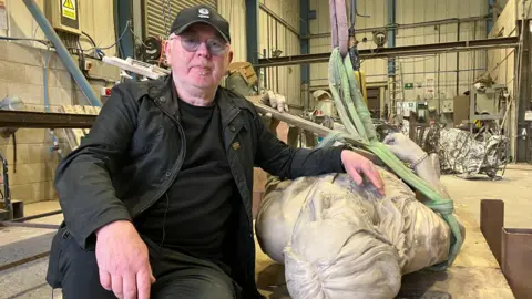 A man with a blonde soul patch and glasses, wearing a black baseball cap, a black t-shirt under a black jacket and black jeans, sits with his hand resting on a statue which is lying on its back.