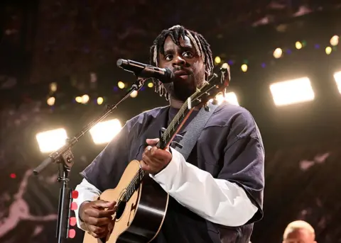 Getty Images Myles Smith performs onstage during iHeartRadio z100's Jingle Ball 2025 in New York City. He holds his guitar while singing pensively.