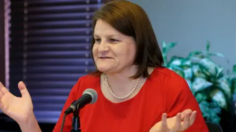 Hannah Dugan gestures while speaking into a microphone in 2016. She is wearing a red top and pearl necklace. She has shoulder-length brown hair.