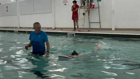 A man is teaching a swimming lesson to a little girl. He is wearing a blue t-shirt and black shorts. He has grey hair. His student is practising kicking her legs. She has her face down in the water and her legs stretched out behind her. She is wearing a turquoise stripey swimming costume. 