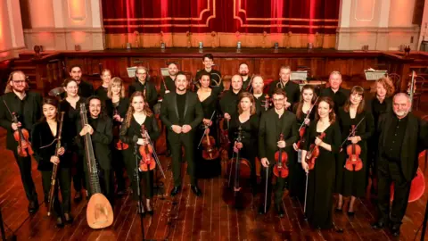 Le Foyer des Artistes A group of international musicians known as Le Foyer des Artistes stand i front of the stage at St Columbs Hall in Derry. They are all dressed in black and holding their instruments
