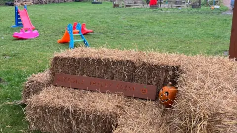 A sign sits on bales of hay, reading Kirkstall Valley Farm next to a pumpkin that has a face carved into it. In the background are two children's slides and two small pieces of play equipment.