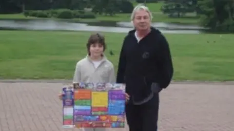 Laura Shirley A grainy picture of Mr Shirley standing next to Laura Shirley in front of a well-kept lawn. He is wearing a black hoodie and trousers and is smiling. Laura is holding a colourful poster while smiling and wearing a cream quarter-zip jumper over a white T-shirt.