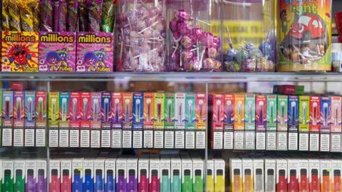 PA Media Disposable vapes of varying colours and flavours on display at a corner shop. There are sweets on display above the vapes.