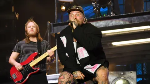 Jim Dyson/Getty Images Teddy Swims performing onstage while singing into a microphone. Next to him is a bass player playing a red bass guitar.