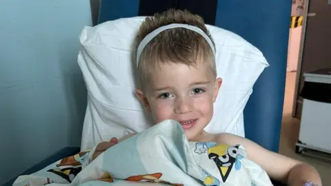 Perry Hill A young boy with blonde hair wearing a white bandage around his head. He is smiling at the camera while sitting on a hospital chair. 