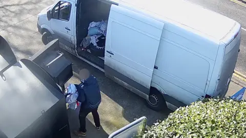 Essex Police A man wearing a navy coat and black trousers holding a large pile of clothes. He is taking it from a large container and is about to load it into his white van, which is parked adjacent to the clothing bank. 