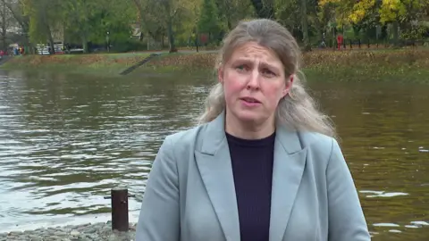 BBC Rachael Maskell has a serious expression as she stands by the River Ouse, a wide waterway in central York. She has long blond hair, tied back and wears a pale green jacked and black T-shirt. Behind her, steep grassy banks, topped by green trees, lead down the the river. 