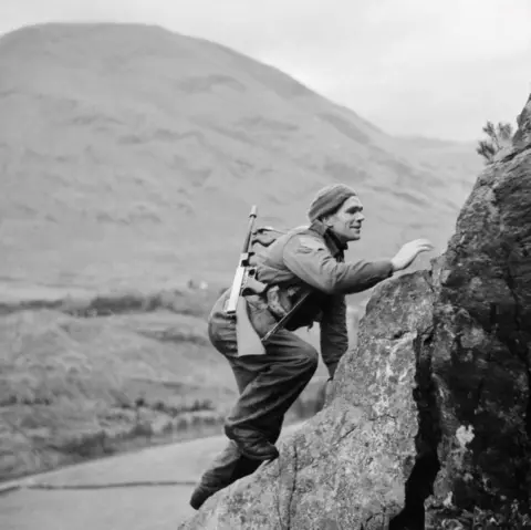 Getty Images In a black and white photograph, a commando wearing combat fatigues climbs a rockface with a machine gun slung over one shoulder. There is a hill in the background.