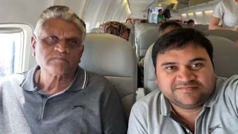 Umang and Kirat Patel take a selfie on board a plane. Both men are wearing grey polo shirts. Umang Patel is smiling slightly while his father is looking quite serious. The seats of the plane are light grey. People can be seen sitting behind Umang and his father and a stewardess is walking up the aisle.