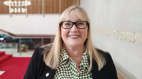 Newcastle City Council Karen Kilgour smiling at the camera. She has medium-length straight blonde hair with a fringe. She is wearing black-rimmed glasses, a green-and-white chequered-patterned shirt and a black suit jacket.