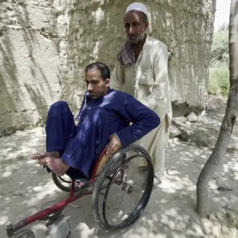 One of the 12 migrants who spoke to BBC Afghan Forensic, an amputee in a blue salwar-kameez, is pushed, in a wheelchair, by another, grey-bearded, man wearing a white one with a white hat and brown patterned scarf, in a tree-lined Afghan landscape.