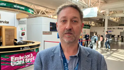 A man wearing a grey suit and blue lanyard standing in an airport departures area with some people in the background.