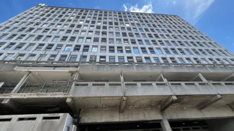 Simon Dedman/BBC A run down concrete office block looking up at it from below. it is a grey building with blue sky above it.