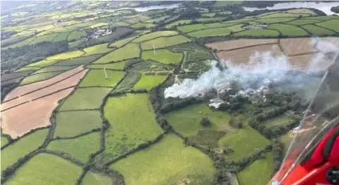 Mike Bawden Aerial image of smoke belching upwards into the atmosphere from a compost fire on the ground which is surrounded by green and verdant arable crop fields and in the distance the gentle bend of a river or lake along with an enticing glimpse of a light plane fuselage painted in a rich and attractive red hue.