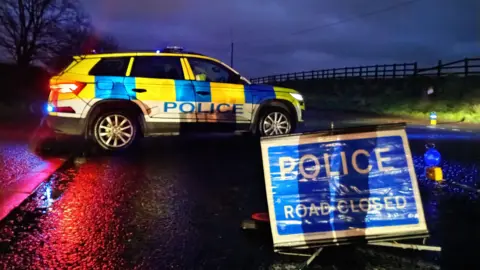 Blue and white police accident sign in front. Behind it is a yellow blue and white police vehicle. It's dark and there is a green bank and fence along the right of the road. 