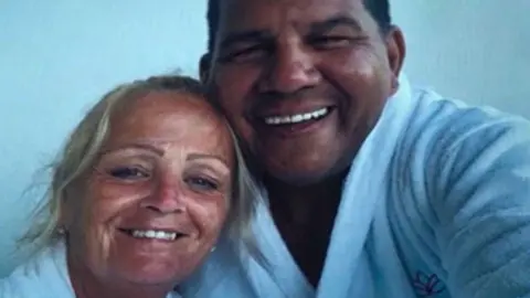 BBC Crimewatch Anita Rose and Richard Jones wearing matching white bathrobes are looking at a camera smiling. Their heads are tilted towards each other.