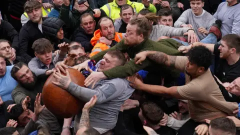 PA Media A group of people, mainly men, tussle for a brown ball. In the centre is a man in a grey top who has the ball clutched to his chest as another man who is horizontal, balanced on top of the crowd, reaches over his shoulders to try to get to the ball. Other men are reaching towards the ball too.