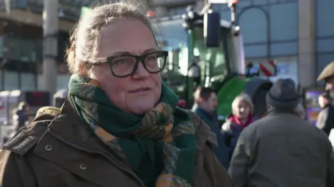 Rachel Hallos stands in front of a tractor in Chester's Town Hall Square. She is wearing a brown coat and green and yellow scarf and is wearing glasses.