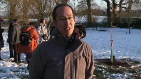 A man wearing a brown coat with dark hair and glasses. Behind him are several other people in coats and snow is on the ground. The newly planted sapling can be seen behind him. 