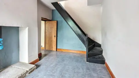 Paul Fosh Auctions A grey tiled floor with a disused fireplace . There's black stairs in the corner. 