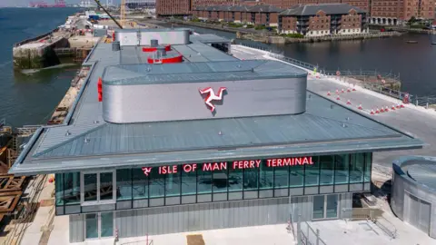 BBC Aerial shot of the front of the ferry terminal, which has Isle of Man Ferry Terminal written across the windows on the second floor and a Manx triskelion on the roof above. There is water either side and red brick building to the right behind it. 