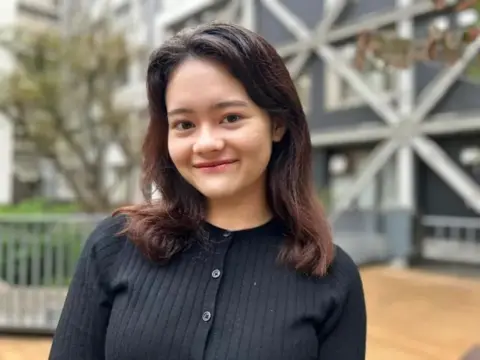 Audrey Hill-Uekawa, 20,  is wearing a black cardigan and has shoulder lenght black hair. She is smiling for the camera standing outside in front of a tree and a building