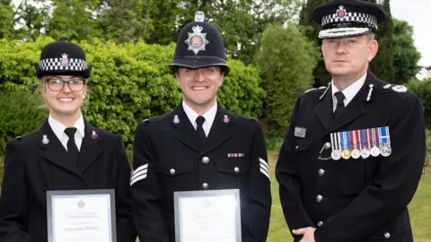 PC Sam Hilbery and Sgt Alex Farr with Essex Chief Constable Ben-Julian Harrington, all in police uniform, facing the camera, they all have police hats on. Sam and Alex are holding certificates in a frame. 