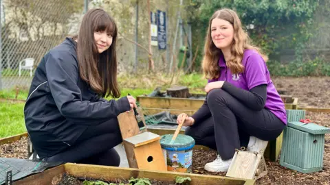 Two female students from Worthing High School are squatting on the ground painting hand-made wooden bird boxes. Both have long hair and are smiling to camera.