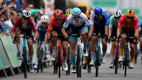  Bradley Collyer/PA Wire Nine cyclists wearing sunglasses and multi coloured outfits and helmets racing with gritted teeth.