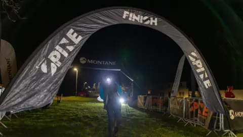 Adam Jacobs Anna Troup coming over the finish line, which is made out of a fabric black arch with the words Spine Finish. Troup's figure is visible but it is dark and her headtorch is lit, meaning her expression is not visible. She is wearing a bright blue outdoor jacket and black trousers.
