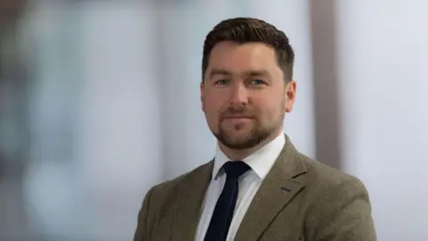 Savills Guernsey A man, with short brown hair, looking at the camera with a blank expression. He is wearing a grey blazer, white shirt and blue tie. 