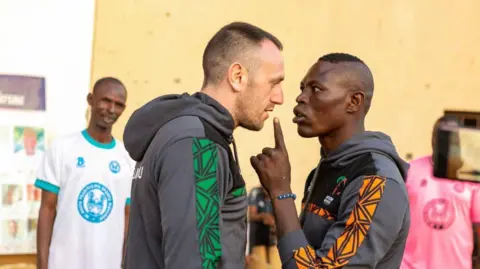 Kayode Oluwa Two men look at one another, standing close together. Both men are wearing black hoodies, one has green down the side of the arm and the other has orange.