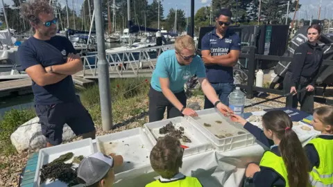 Two men in blue t-shirts stand either side of another man wearing a light blue top, who is leaning over a table on top of which are several plastic trays containing small items. He is handing an item to a young girl who is sitting the other side of the table in between other children. In the background is a marina with several moored boats and tall trees behind those.
