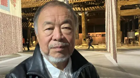 Photograph of the artist Ai Weiwei. He is pictured alongside his artwork in the giant warehouse space at Manchester's Aviva Studios. 